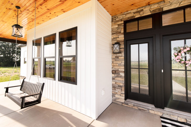 A modern farmhouse featuring a front entry with double black doors, a wooden ceiling, stone facade, and a spacious sitting porch.