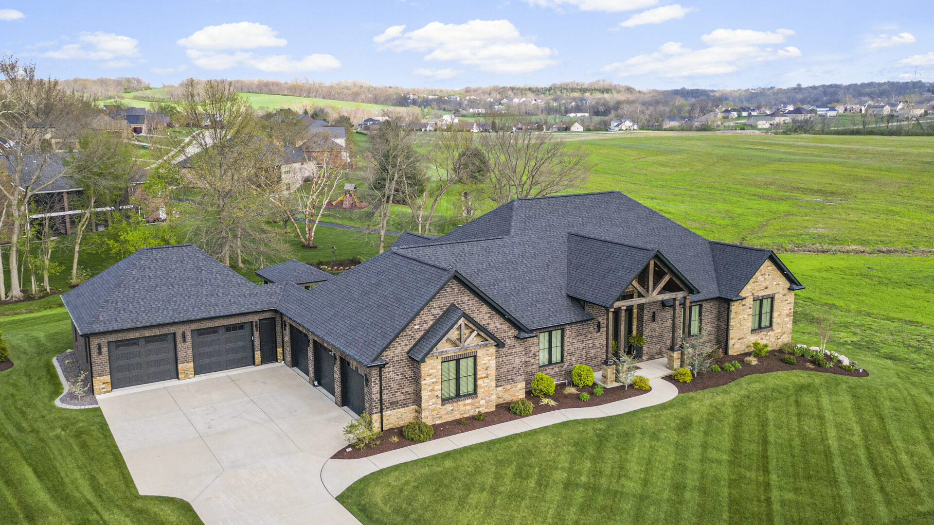 An aerial drone captures a contemporary mountain-style home, showcasing its stone and brick exterior. Dark wooden beams extend over the front entrance, lending an air of grandeur.