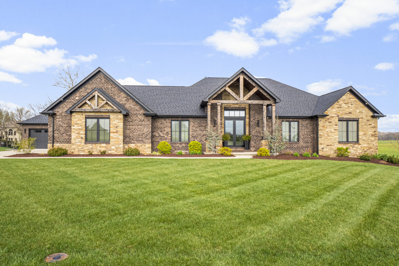 A contemporary mountain-style home features a stone and brick exterior. Over the front entrance, dark wooden beams stretch out, adding a touch of grandeur.